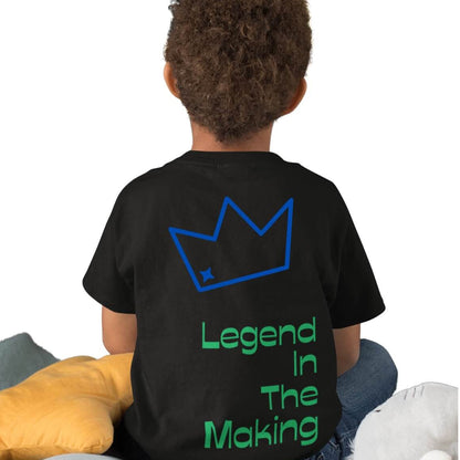 Back view of a biracial child wearing a black t-shirt with a blue crown and green text 'Legend In The Making' on the back, sitting on a bed with pillows. 