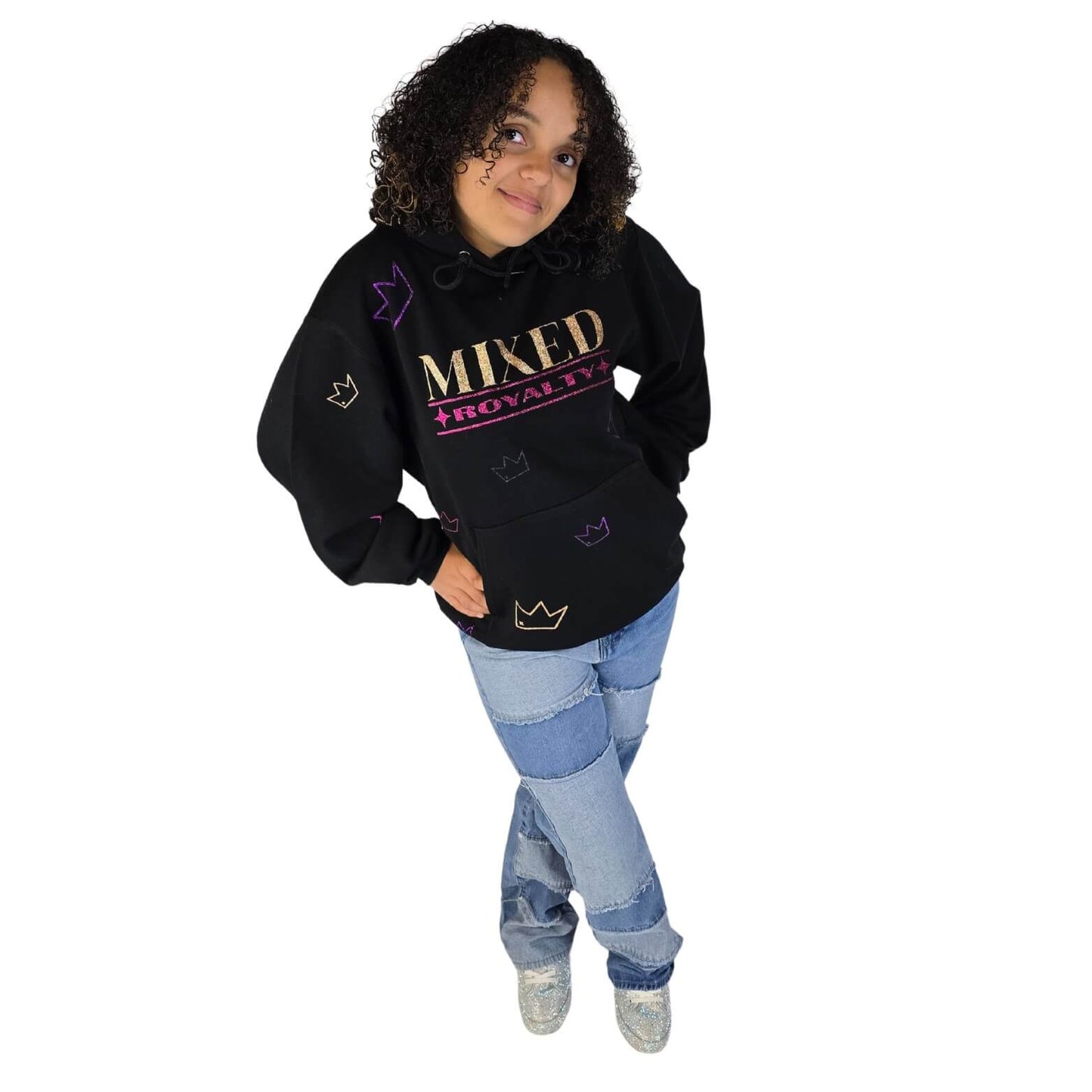 Smiling mixed-race girl wearing a black 'Mixed Royalty' hoodie with gold and purple sparkle lettering and sparkle crowns randomly all over with patchwork jeans, and sparkly sneakers.  Youth empowerment fashion promoting biracial pride.