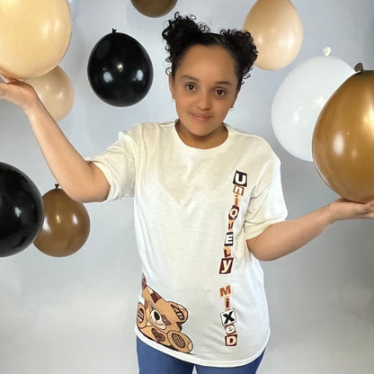 Smiling child wearing Uniquely Mixed white t-shirt with vertical logo and teddy bear graphic, surrounded by black, brown, and white balloons, representing inclusive kids fashion and playful streetwear style.
