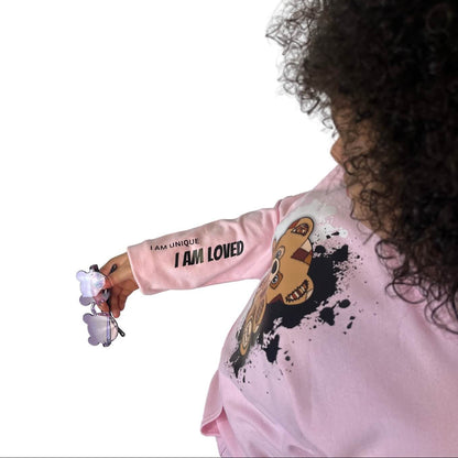 Biracial child wearing a pink sweatshirt with text "I Am Unique I Am Loved" on the sleeve and graphics, holding teddy bear shaded glasses. Fun and cute affirmation inspired kids clothing. 